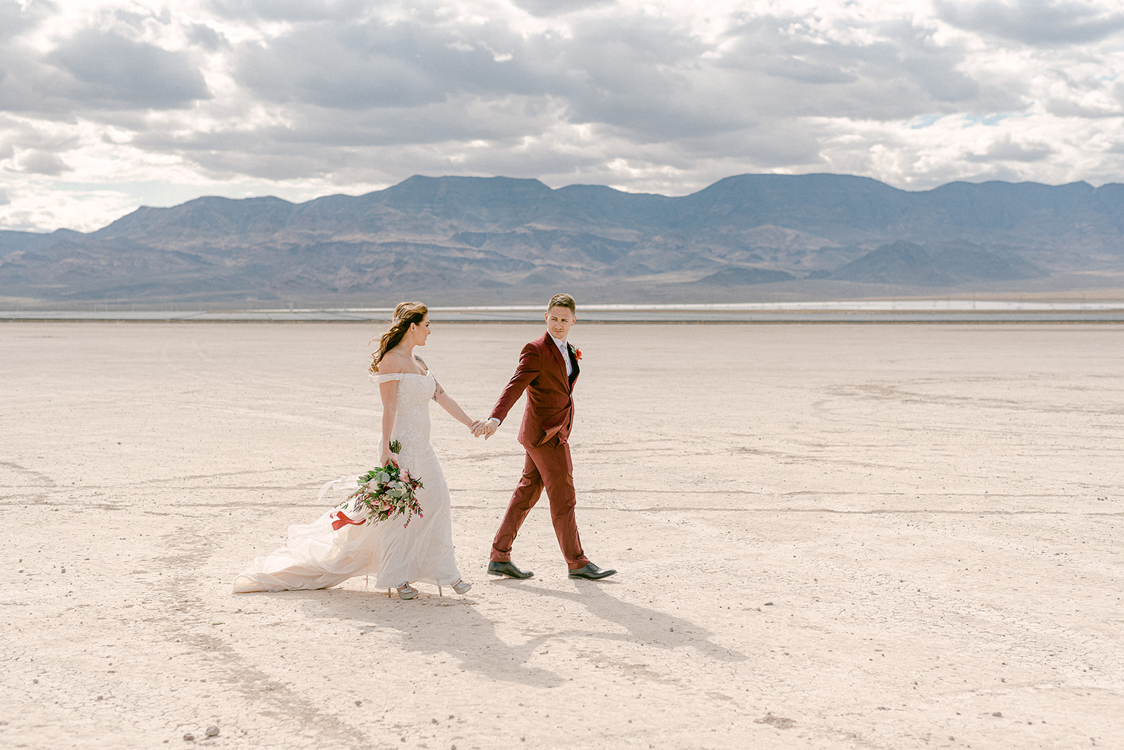 Groom looks back at the bride as they hold hands and walk along Dry Lake Bed, photographed by Emily Reno