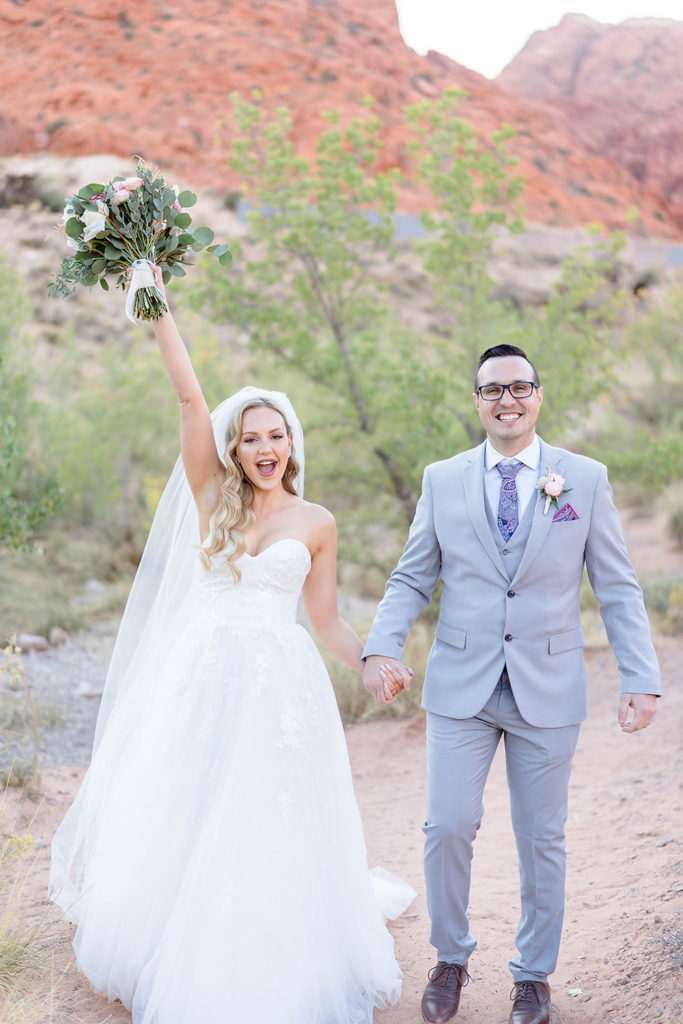 Red Rock Ash Springs: Bride raises her bouquet as she holds hands with the groom as they smile for the camera