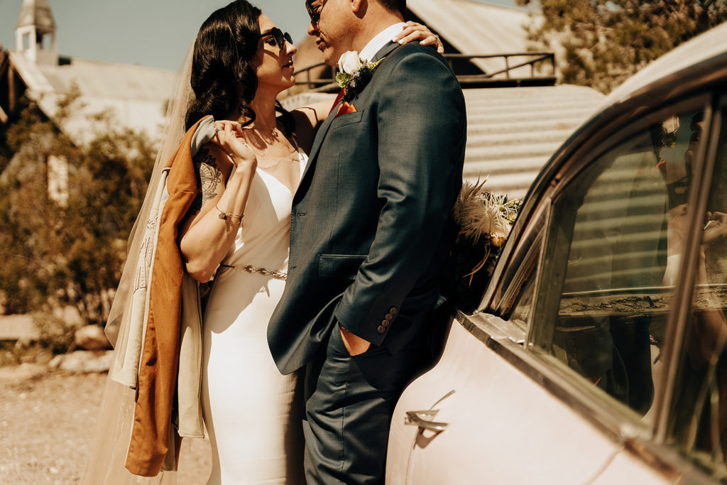 Eloping in Vegas: Ultimate Guide (2022). Bride and groom sharing an embrace as they lean against a car with sunglasses on.