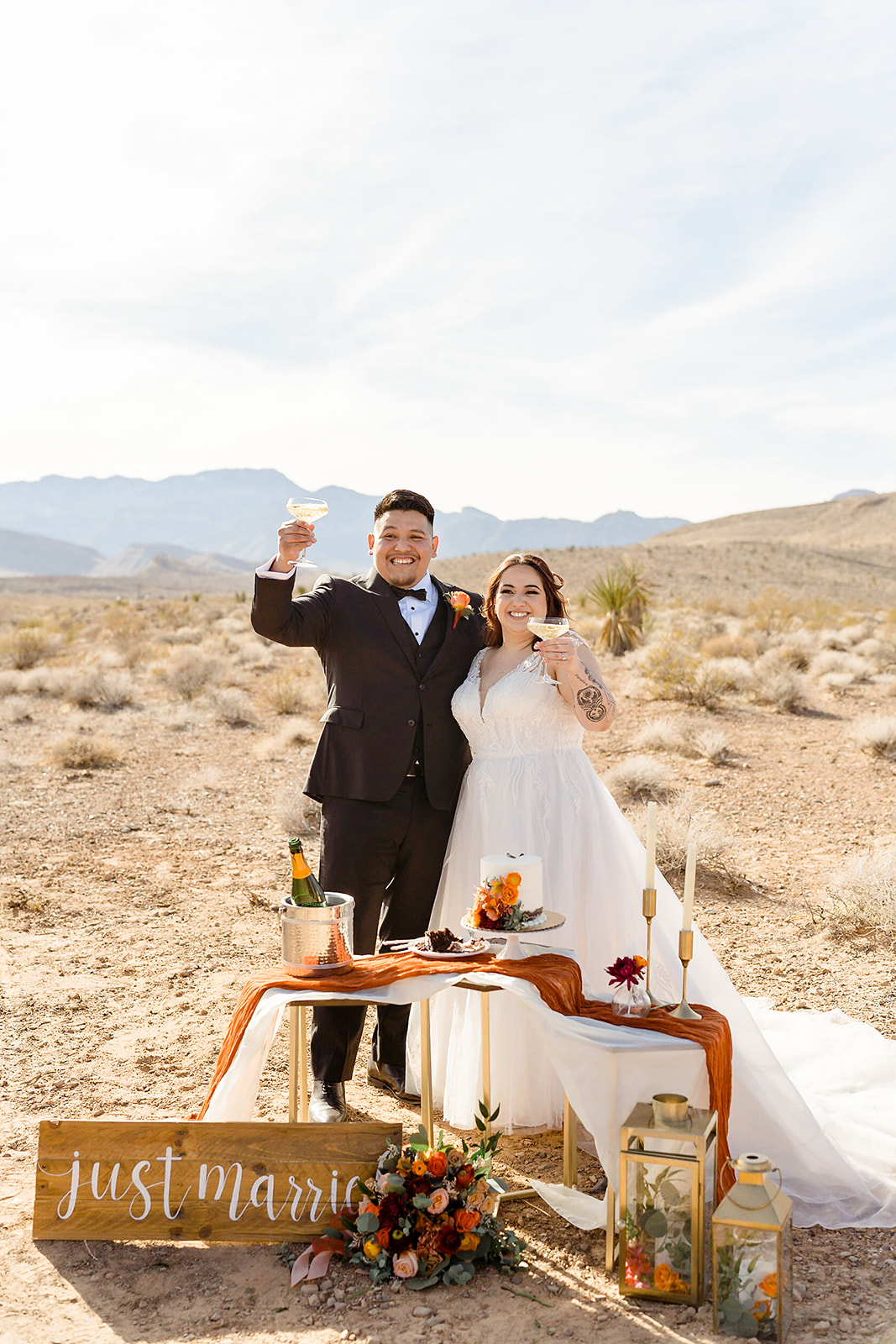 Bride and groom smile as they raise their glasses during their elopement shoot in Las Vegas with their wedding cake and a sign that reads Just Married in front of them