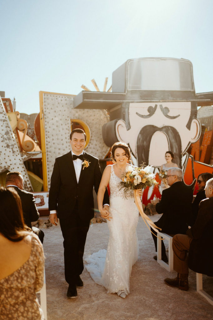 Bride and groom smiling as they hold hands while walking back down the aisle during their wedding at Neon Museum