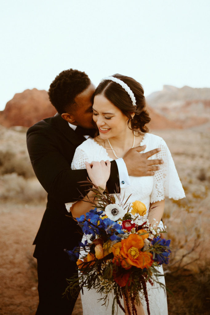 Groom hugs bride from behind as she smiles and holds her vibrant bouquet during their wedding shoot with Elopement Las Vegas