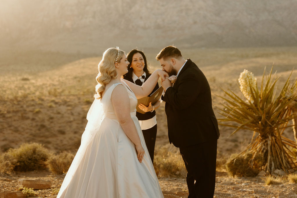 Groom plants a kiss on bride's hand as she and the officiant smile during their wedding arranged by Elopement Las Vegas