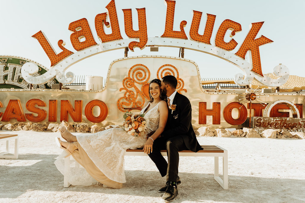 Neon Museum Wedding & Elopement Package. Couple smiling as they sit on bench with signs behind them.