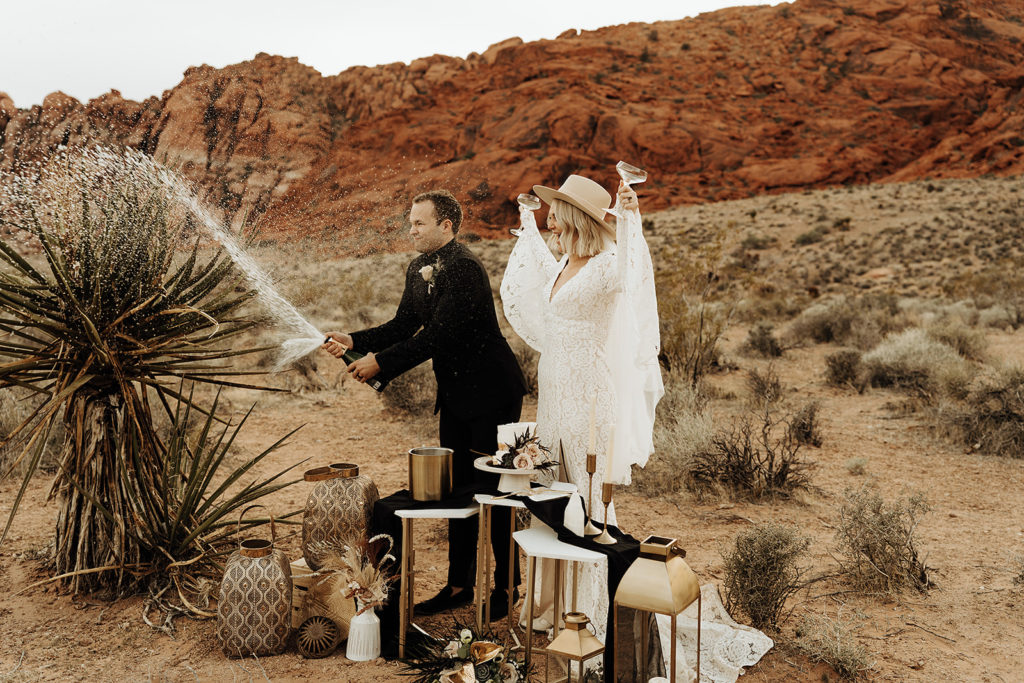 Groom spraying champagne as bride holds up two glasses during their outdoor Las Vegas elopement