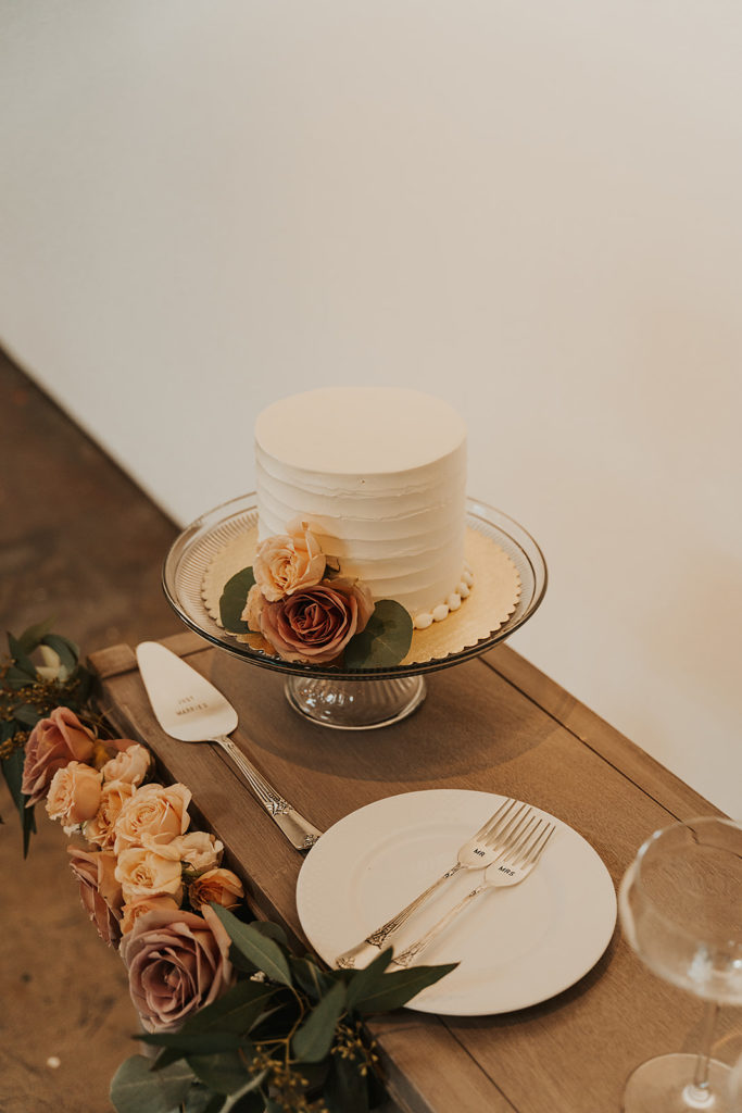 Simple vanilla cake with rose decor placed on wooden table with knife, plates, forks and glasses