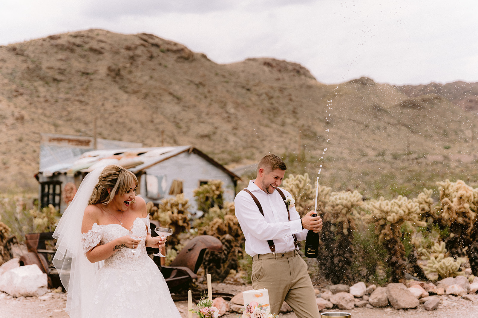 Groom popping open a bottle of champagne as bride looks on during their Nelson Ghost Town wedding reception planned by Elopement Las Vegas