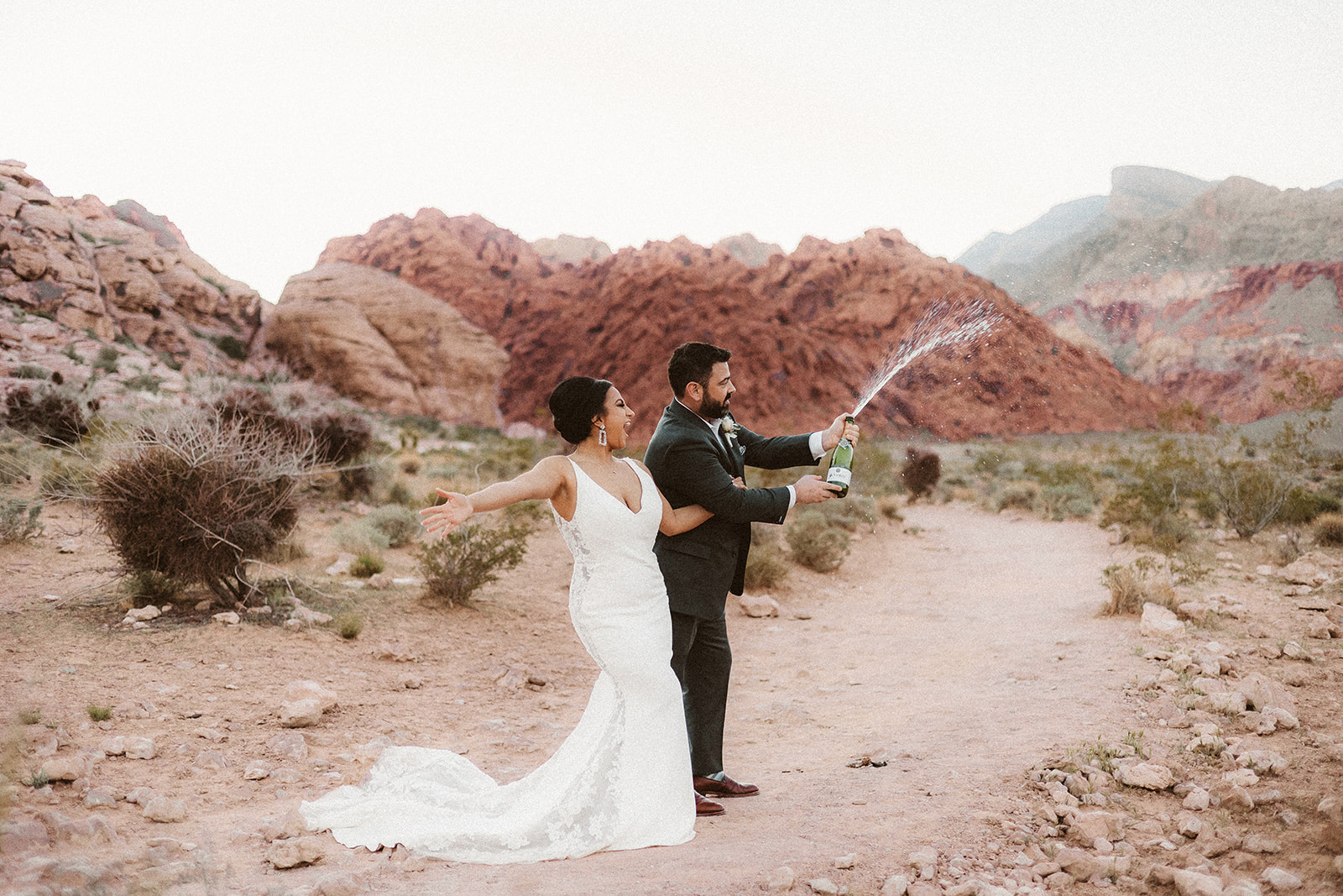 Bride cheers on groom as he pops open a bottle of champagne during wedding planned by Elopement Las Vegas