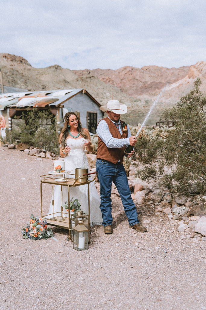 Bride smiles as she watches groom pop open a bottle of champagne during reception planned by Elopement Las Vegas