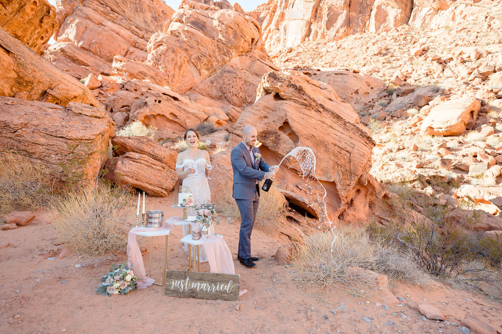 Groom pops open a bottle of champagne as bride holds glasses beside him during their Las Vegas elopement