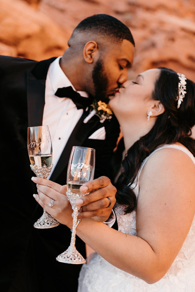 Bride and groom sharing a kiss as they hold champagne glasses during their elopement coordinated by Elopement Las Vegas