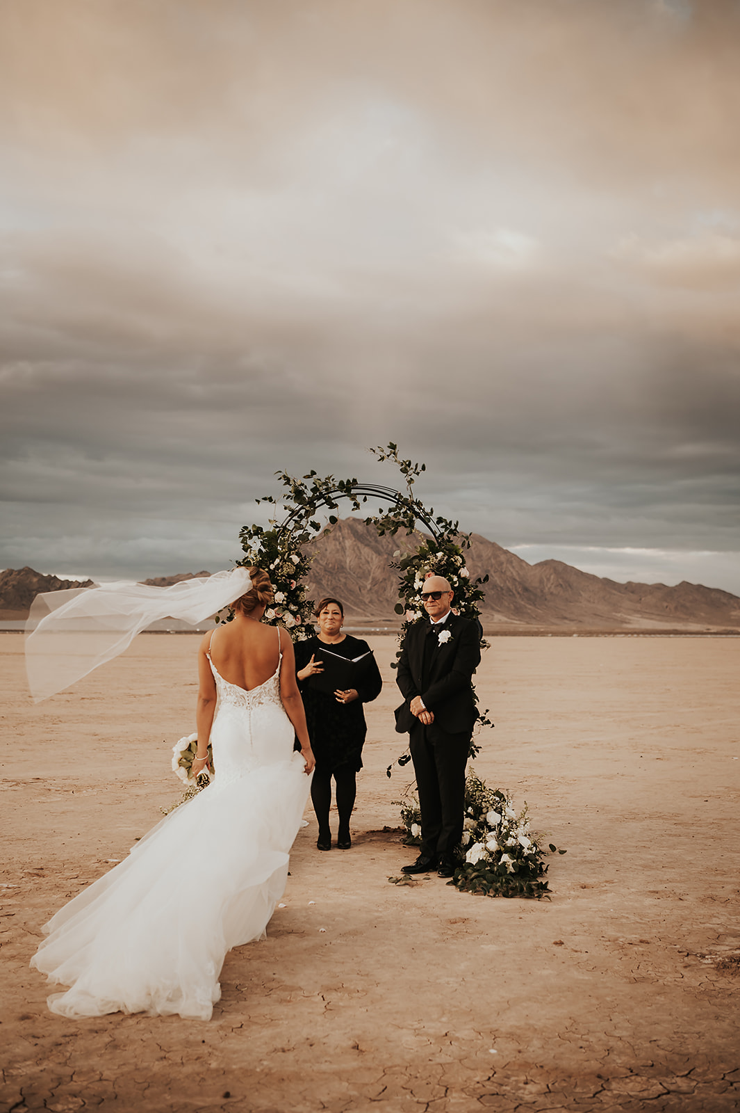 Dry Lake Bed Wedding & Elopement Guide. Bride walking towards the wedding arch with groom and officiant smiling at her.