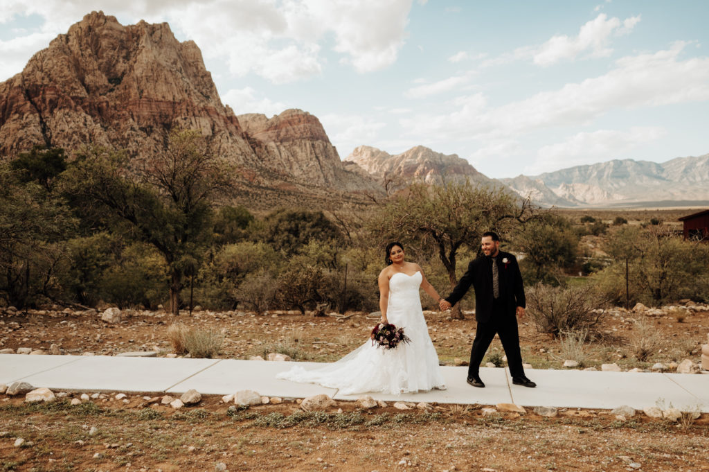 Newlywed couple holding hands and walking with blue skies and mountains behind them